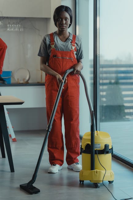 A team of three professional cleaners, dressed in matching grey shirts and red overalls, standing in a modern living room with light-colored walls, performing surface cleaning tasks. The room features