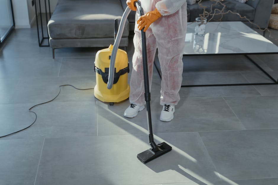 A professional cleaner, a young woman with dark hair tied back, wearing a black cleaning uniform over a white shirt, standing indoors against a plain light-colored wall. She holds a white spray bottle